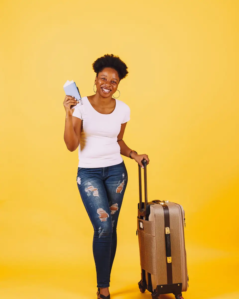 A smiling young woman holds her passport and her suitcase.